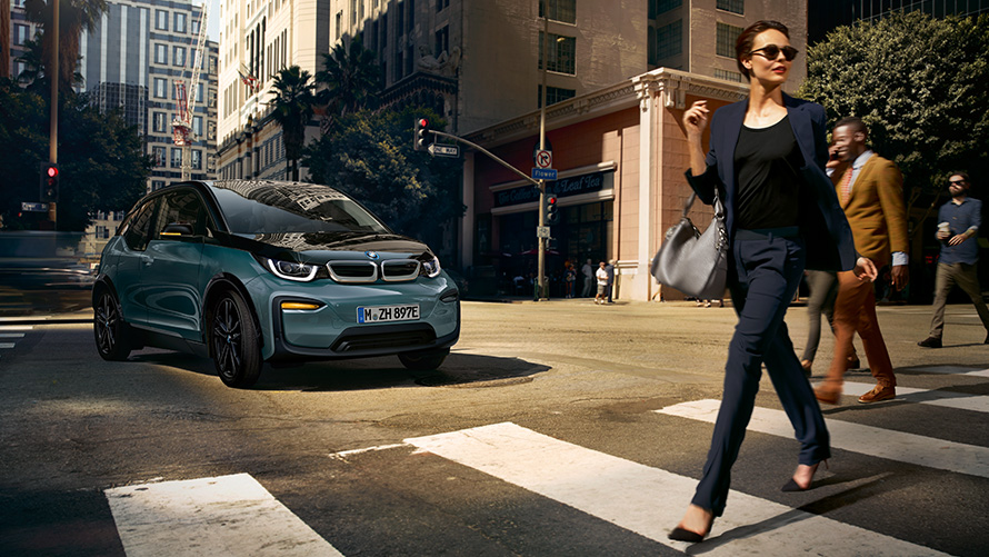 BMW i3 I01 Blue Ridge Mountain metallic mit Akzent Frozen Grey metallic Dreiviertel-Frontansicht mit Frau über Zebrastreifen laufend