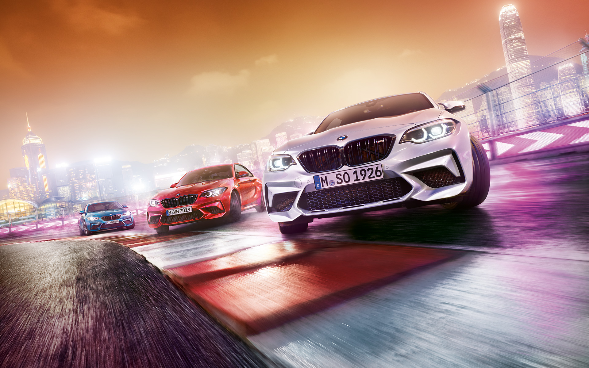 BMW M2 Competition in Aktion Drei BMW M2 Competition driften auf Rennstrecke vor nächtlicher Großstadtkulisse.