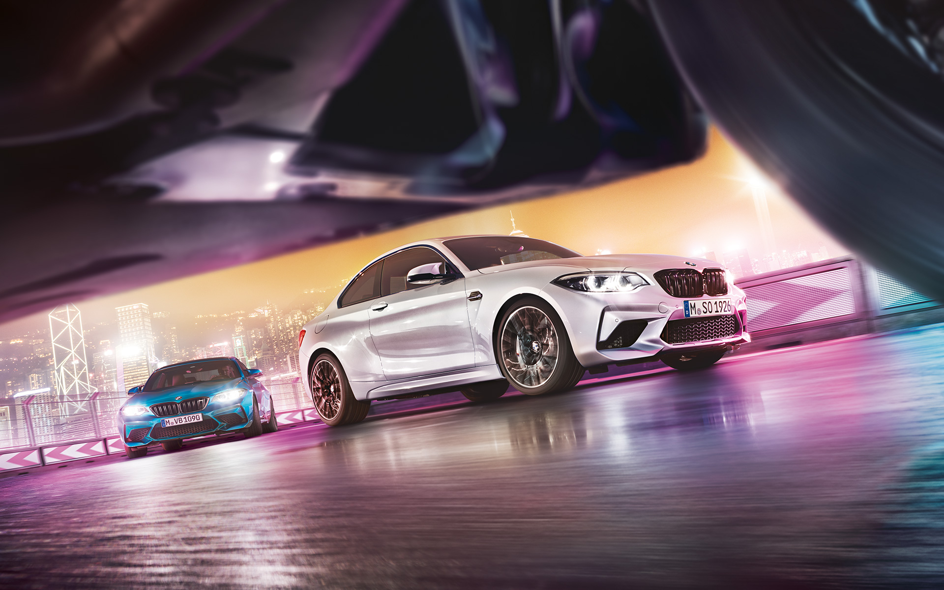 BMW M2 Competition auf spiegelndem Asphalt Zwei BMW M2 Competition auf Rennstrecke vor nächtlicher Großstadtkulisse aus Bodenperspektive.
