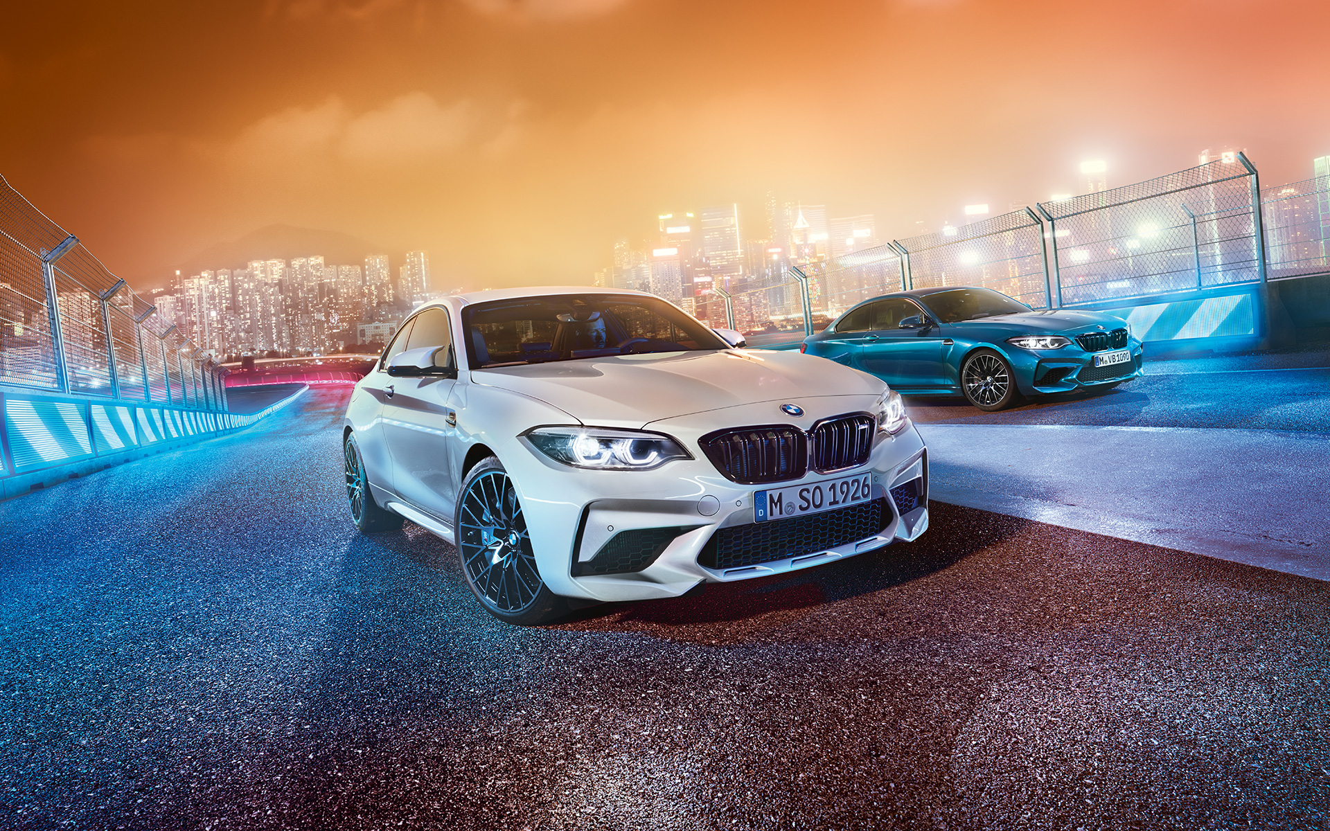 Zwei BMW M2 Competition auf der Rennstrecke Weißer und blauer BMW M2 Competition in schräger Frontansicht stehend vor nächtlicher Großstadtkulisse.