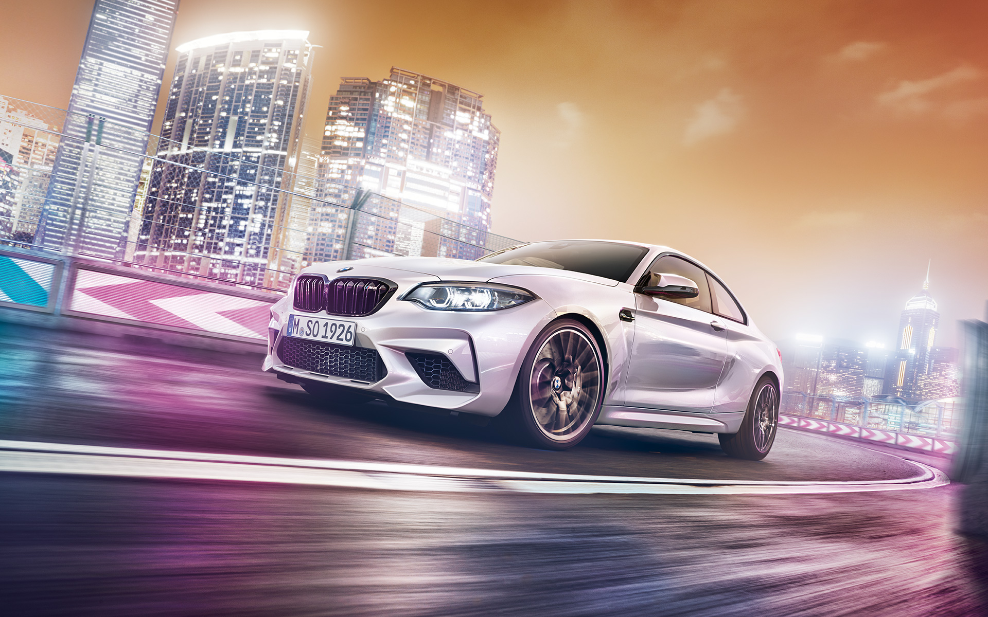 Weißer BMW M2 Competition mit Skyline Weißer BMW M2 Competition fährt auf Rennstrecke vor nächtlicher Skyline.