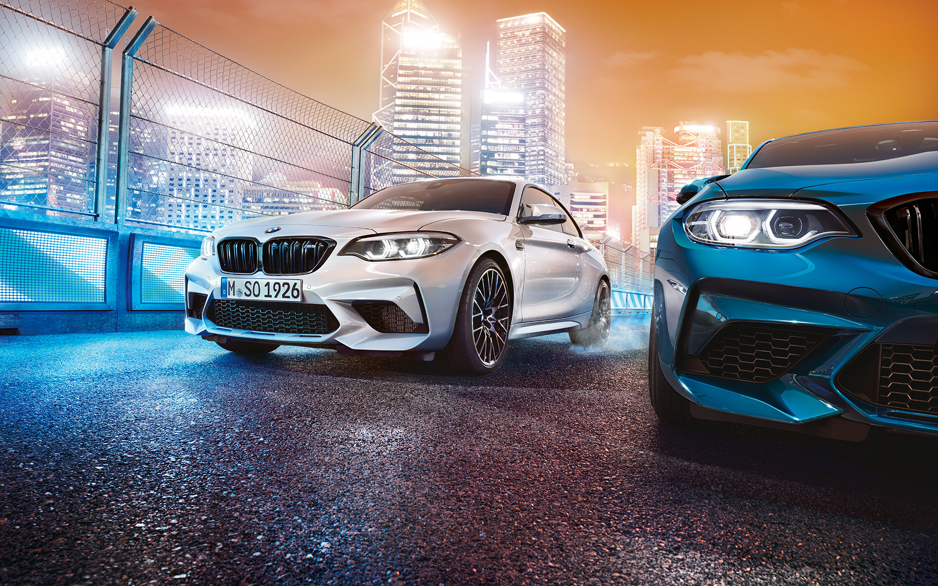 Weißer und blauer BMW M2 Competition im Stand Weißer und blauer BMW M2 Competition in der Frontansicht stehen vor nächtlicher Großstadtkulisse.