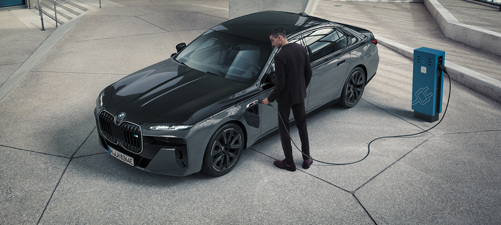 BMW M760e xDrive Limousine G70 M Performance Paket BMW BMW Individual Two-Tone Lackierung Saphirschwarz metallic BMW Individual Dravitgrau metallic Dreiviertel Frontansicht Mann mit Ladekabel neben Ladestation