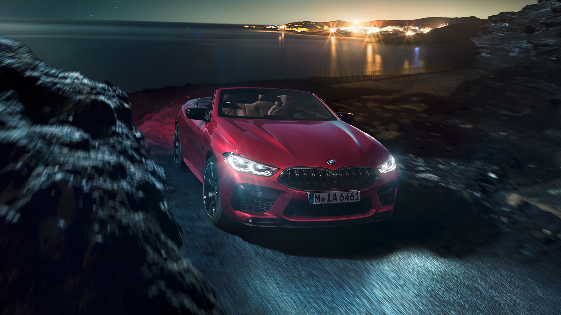 BMW M8 Competition Cabriolet, Dreiviertel-Frontansicht. BMW M8 Competition Cabriolet in Motegi Rot metallic mit geöffnetem Verdeck, Dreiviertel-Frontansicht, fahrend bei Nacht auf Küstenstraße.