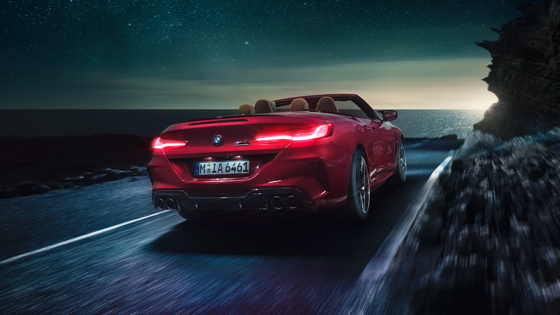 BMW M8 Competition Cabriolet, Dreiviertel-Heckansicht. BMW M8 Competition Cabriolet in Motegi Rot metallic mit geöffnetem Verdeck, Dreiviertel-Heckansicht, fahrend bei Nacht auf Küstenstraße.