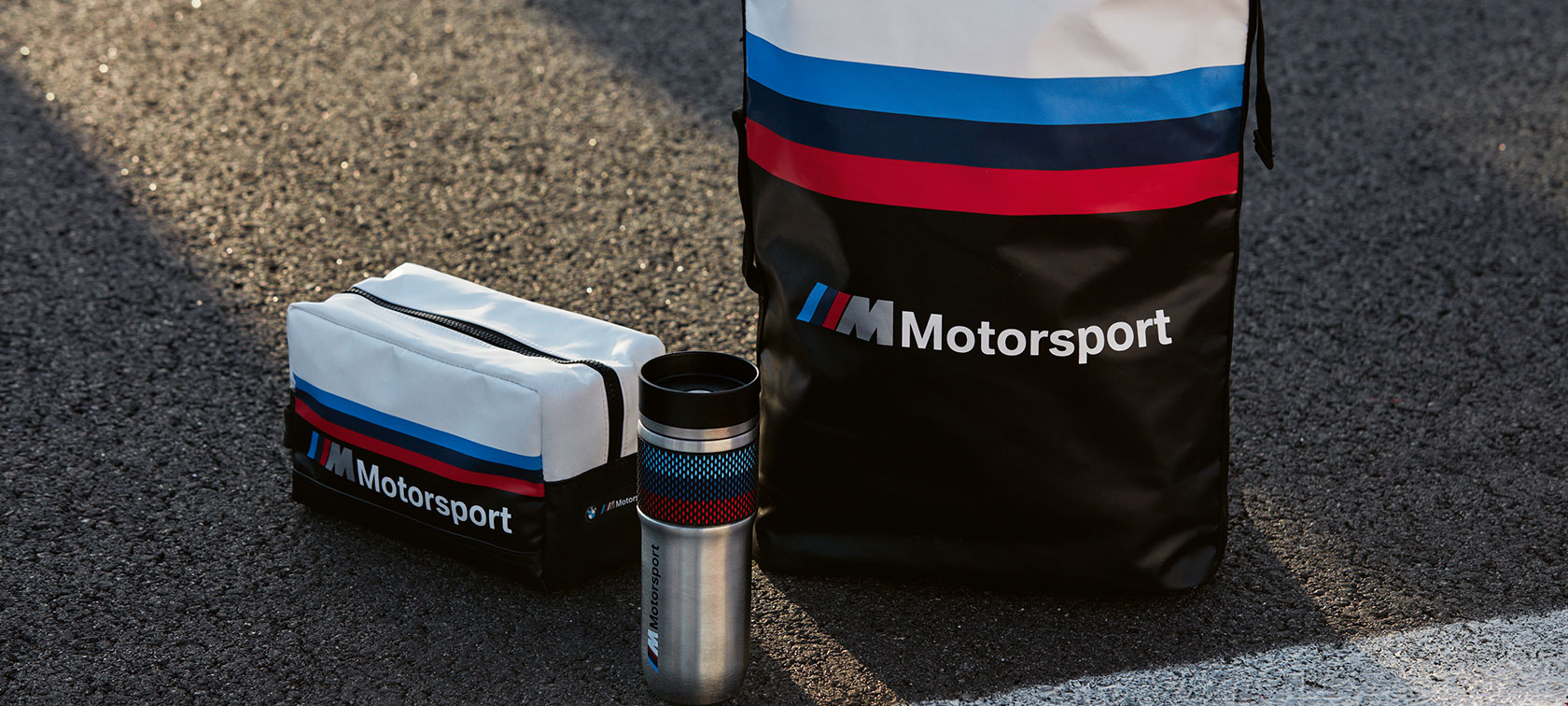 Auf dem Bild stehen Kulturbeutel, Thermobecher und Rucksack aus der BMW M Motorsport Collection nebeneinander. Auf dem Bild stehen Kulturbeutel, Thermobecher und Rucksack aus der BMW M Motorsport Collection nebeneinander.