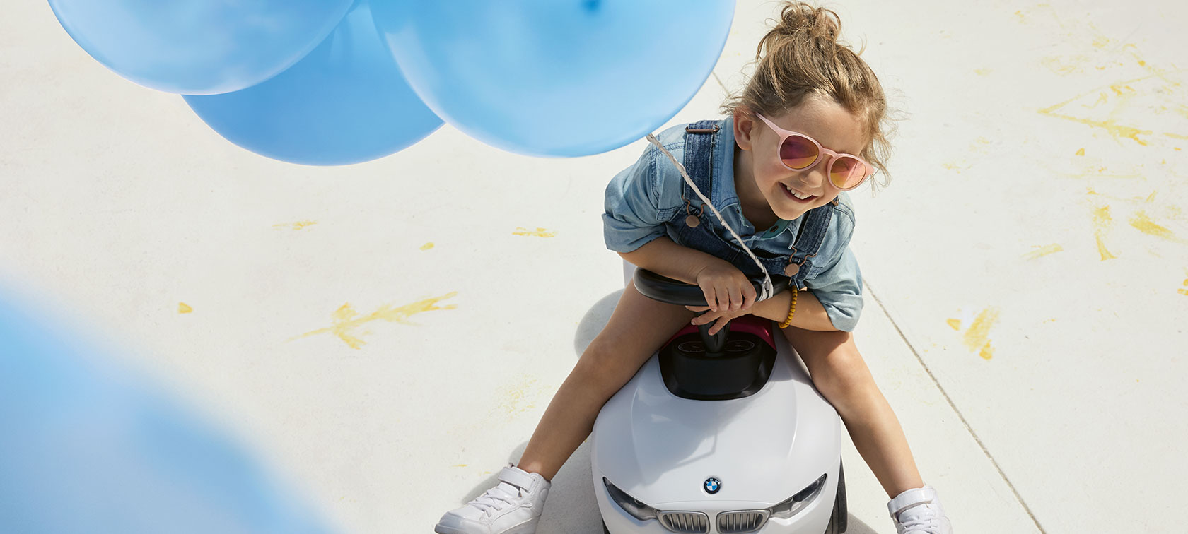 BMW Kids. Ein Mädchen mit rosa Sonnenbrille und einem blauen Ballon in der Hand sitzt fröhlich auf einem BMW Babyracer.