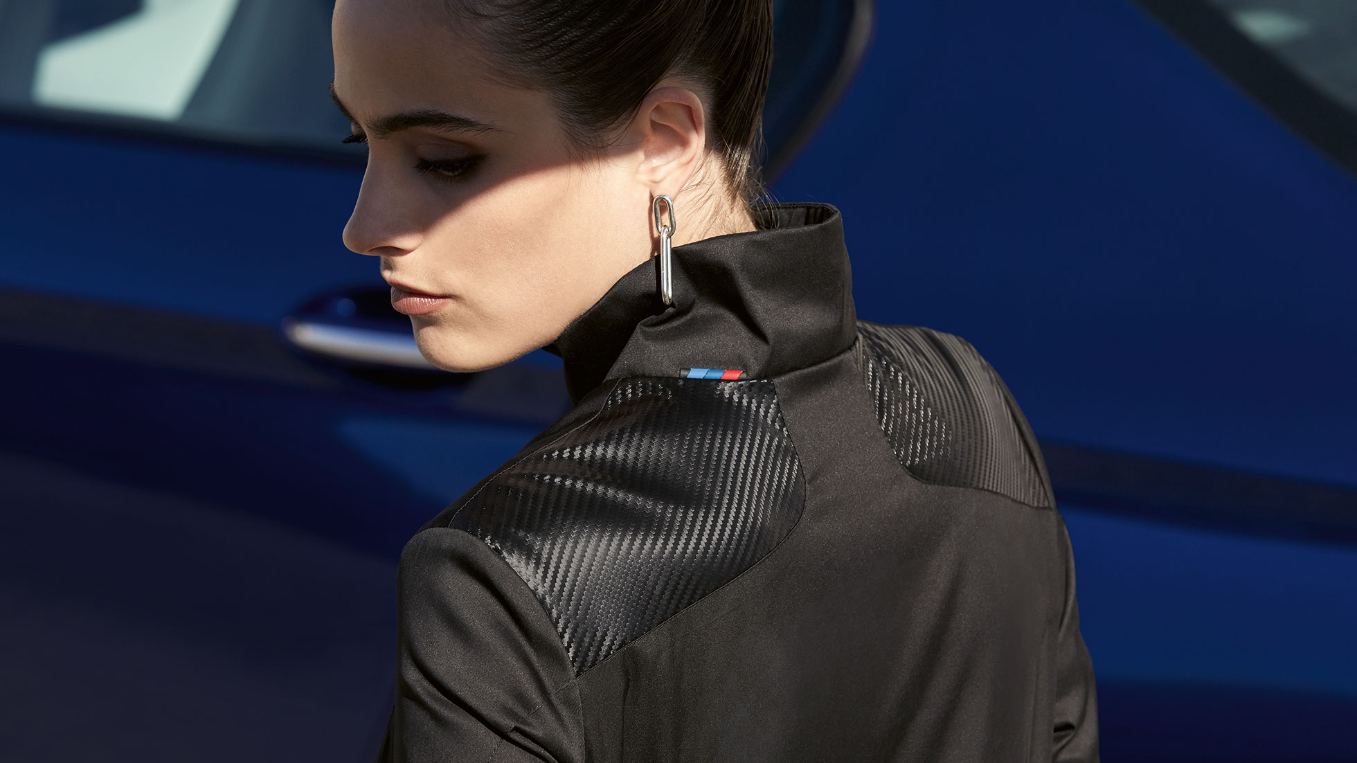 BMW M Jacke Damen. Ein weibliches Model blickt über ihre Schulter und trägt dabei die BMW M Jacke für Damen.