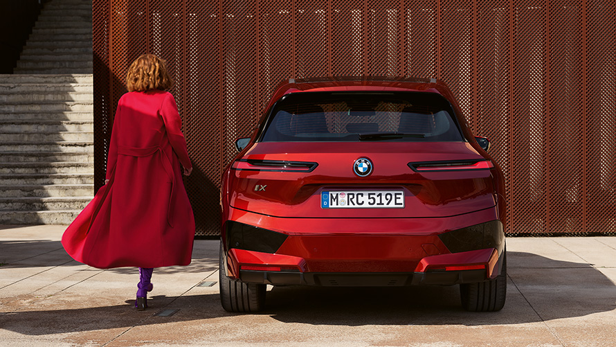 Frau in rotem Mantel neben BMW iX (i20) Schließen der Heckklappe Schließen der Heckklappe Frau in rotem Mantel neben BMW iX (i20) vor Gebäude