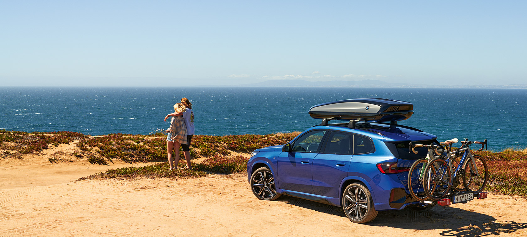 Original BMW Zubehör für Frühling und Sommer BMW X1 U11 Original BMW Zubehör für Frühling und Sommer BMW X1 U11 auf Sand stehend vor einer Küste mit 2 Personen